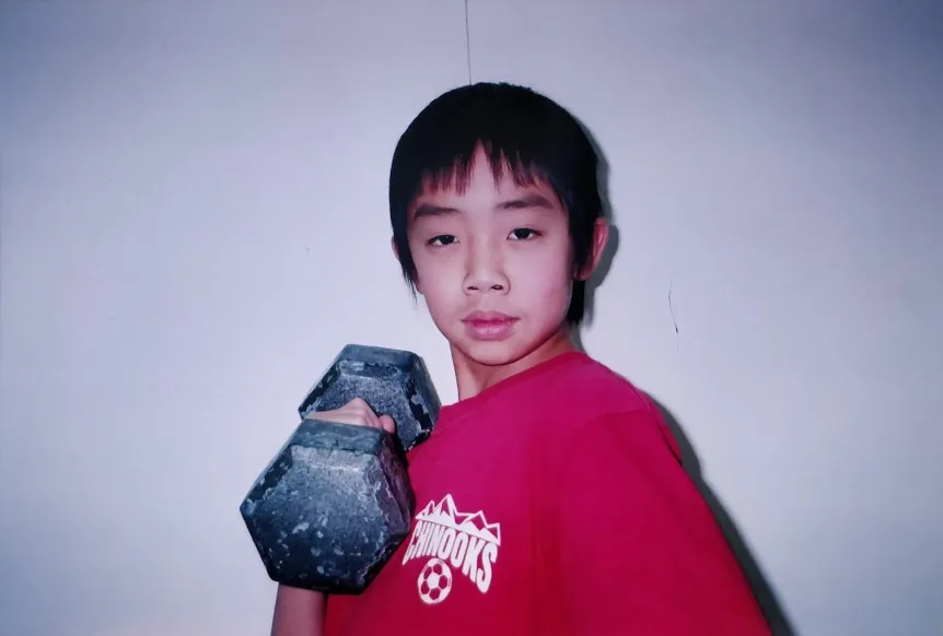 Preteen Brennan in a red Chinooks soccer shirt holding a dumbbell up to his shoulder, looking seriously at the camera.
