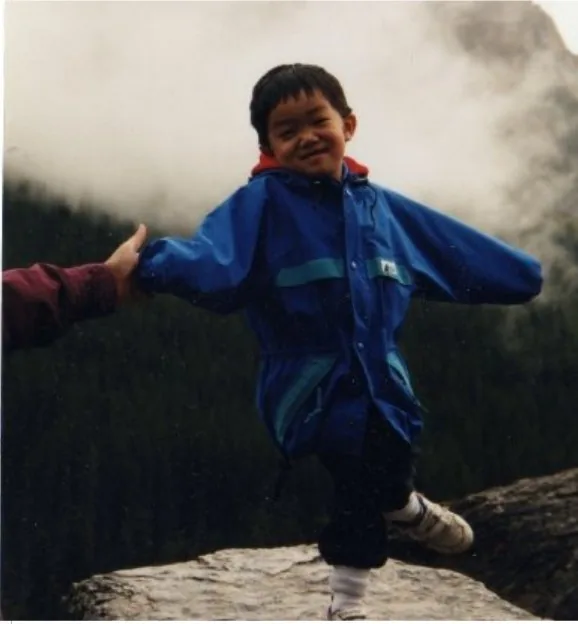 Young Brennan in a blue rain jacket balancing on one leg on a rock, arms out and grinning, with misty mountains behind him.