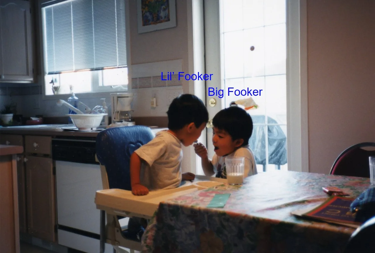 Old family photo of two young brothers at a kitchen table: Brennan, labelled “Lil’ Fooker,” sits in a high chair while Jordan, labelled “Big Fooker,” feeds him a spoonful.