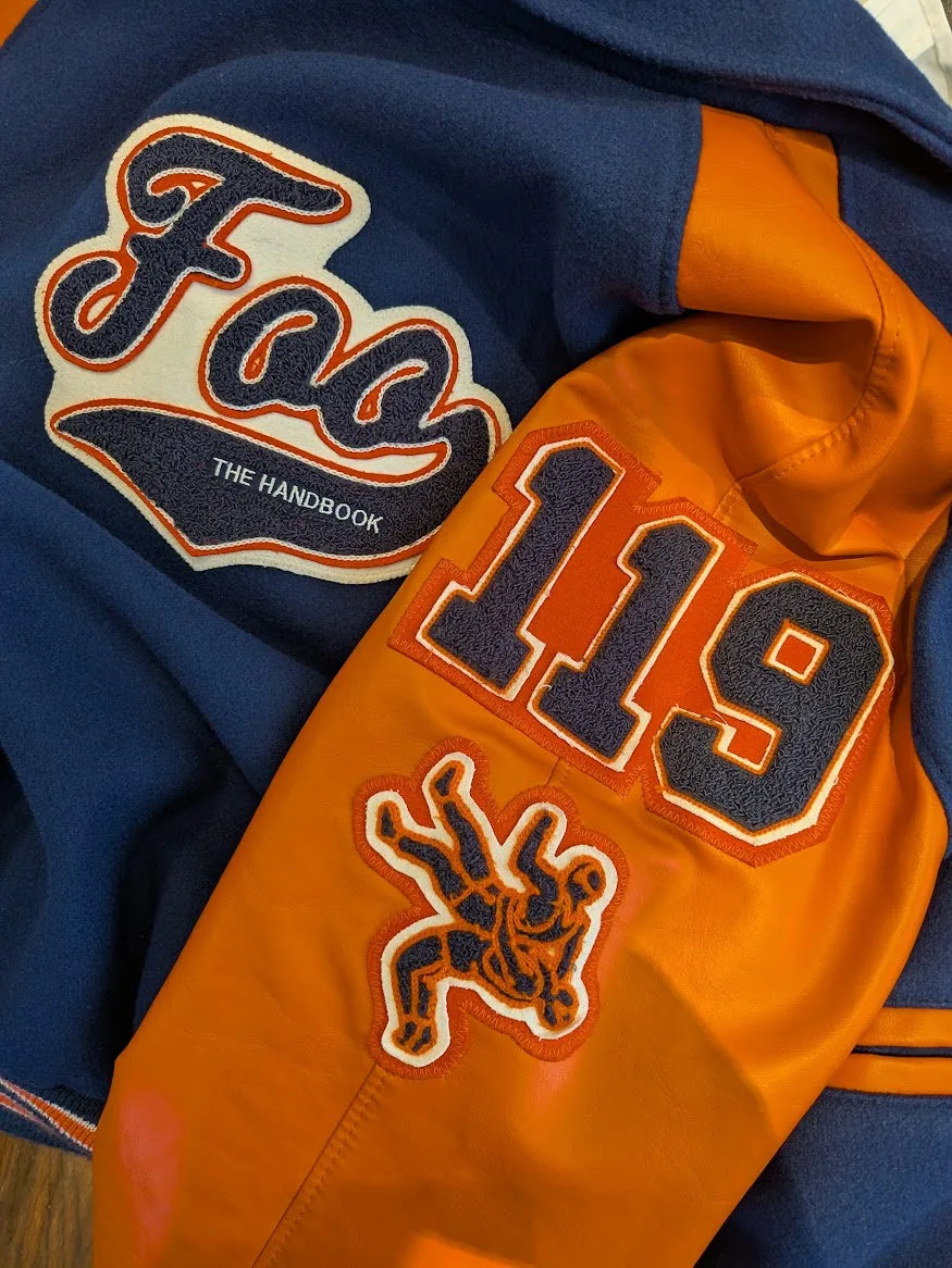 Orange and blue letter jacket with a “Foo” patch reading “The Handbook,” the number 119, and a wrestling patch on the sleeve.