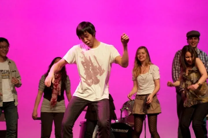 Brennan in a white T-shirt dancing mid-performance at the front of a stage, with other singers laughing and dancing behind him against a bright pink backdrop.